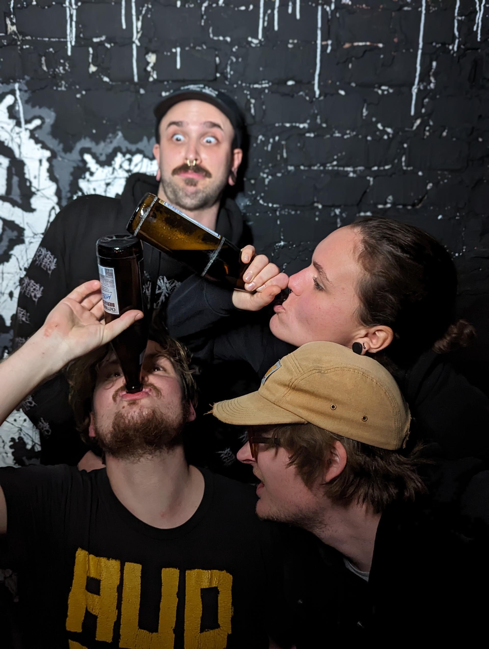 4 Bandmitglieder vor einer schwarzen Wand. Zwei von vier trinken ein Bier. Die anderen zwei schauen doof aus der Wäsche.