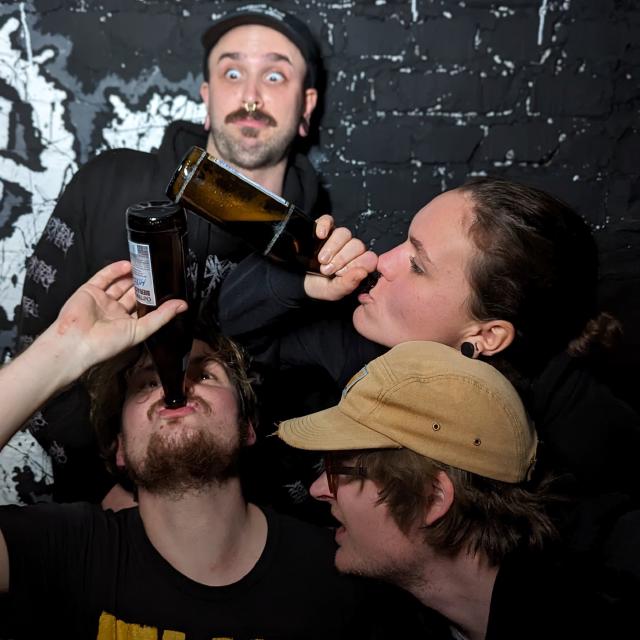 4 Bandmitglieder vor einer schwarzen Wand. Zwei von vier trinken ein Bier. Die anderen zwei schauen doof aus der Wäsche.
