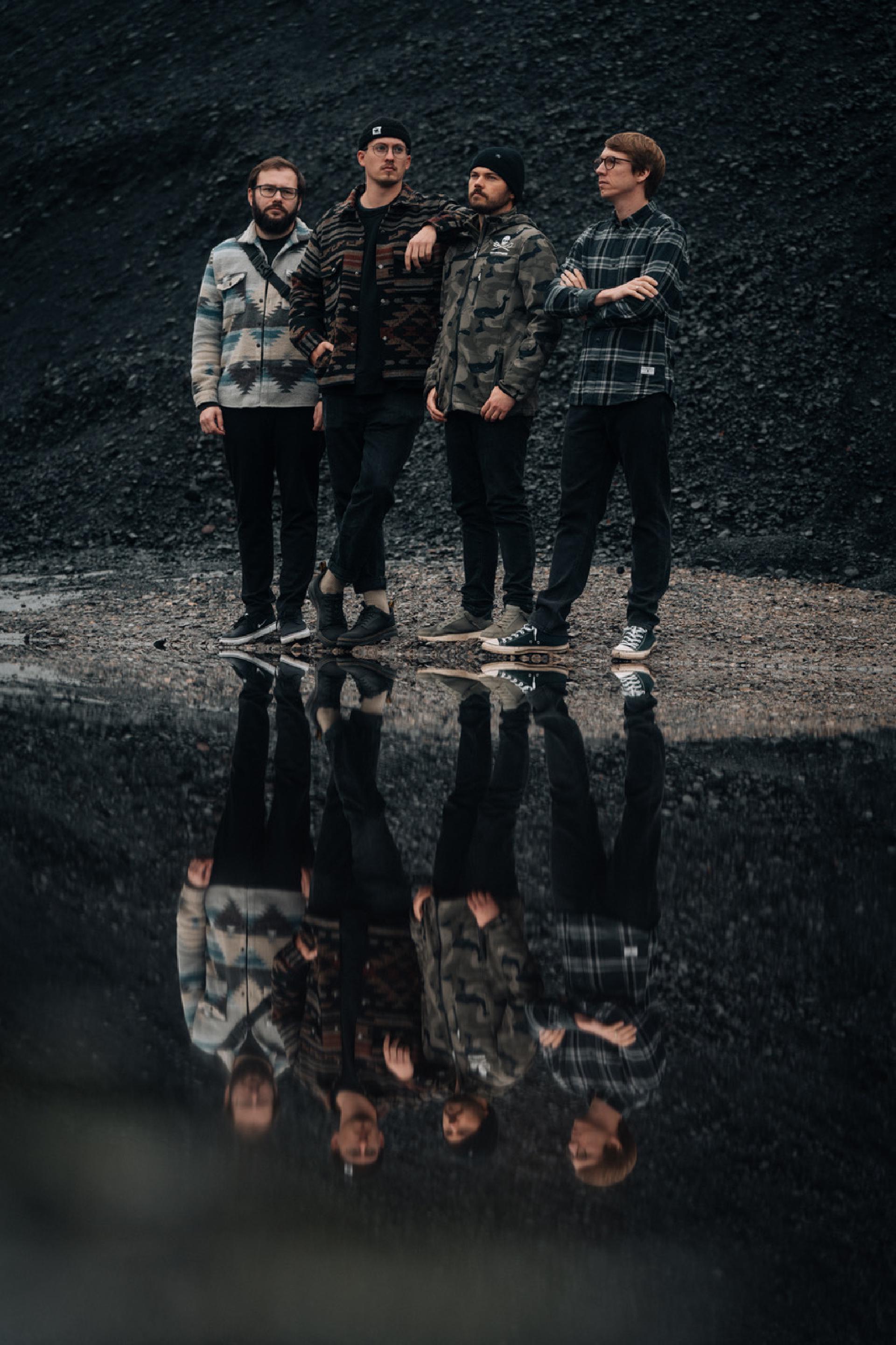 Band Devil May Care steht im Regen zusammen auf einem spiegelnden Untergrund, sodass sie auf dem Boden gespiegelt dargestellt werden
