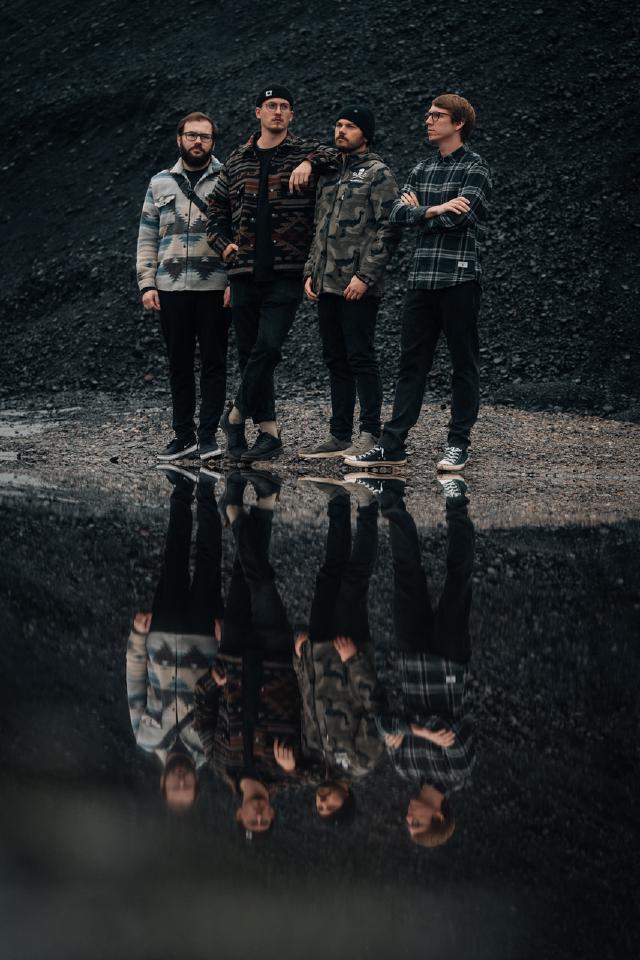 Band Devil May Care steht im Regen zusammen auf einem spiegelnden Untergrund, sodass sie auf dem Boden gespiegelt dargestellt werden